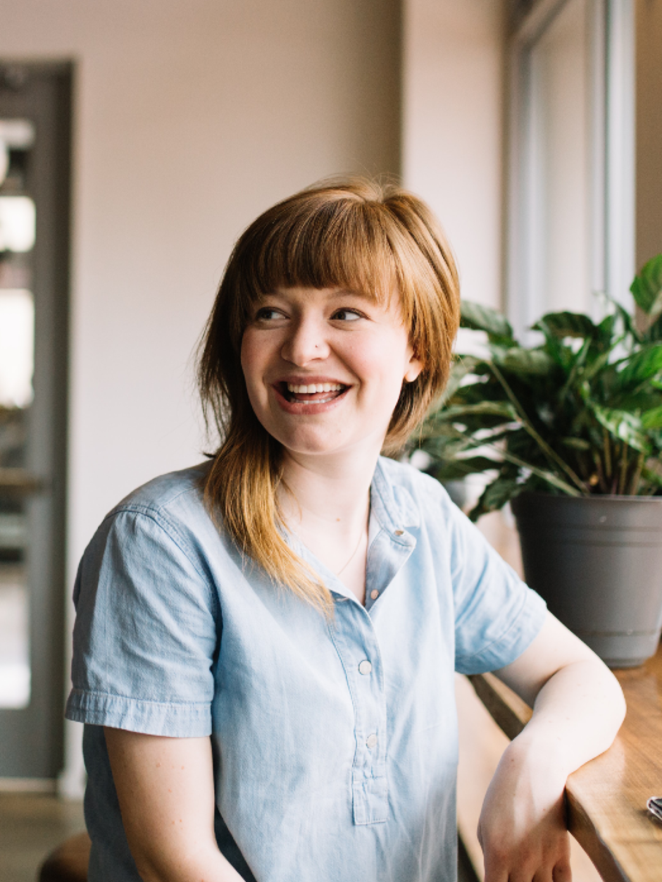 A smiling lady looking away out of shot smiling with a green plant behind her