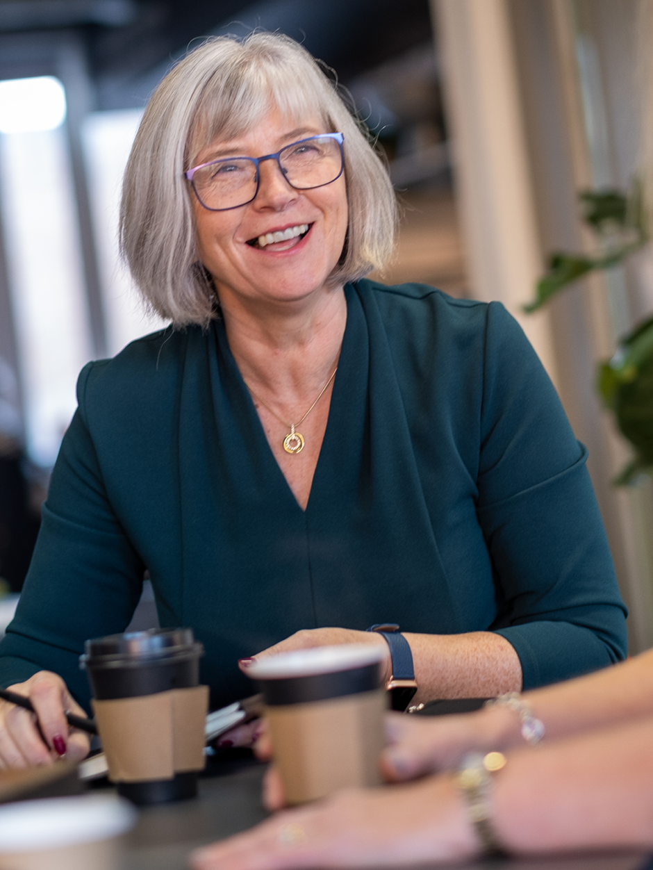 A lady in glasses and a green dress with a coffee, smiling at the person out of shot across the desk